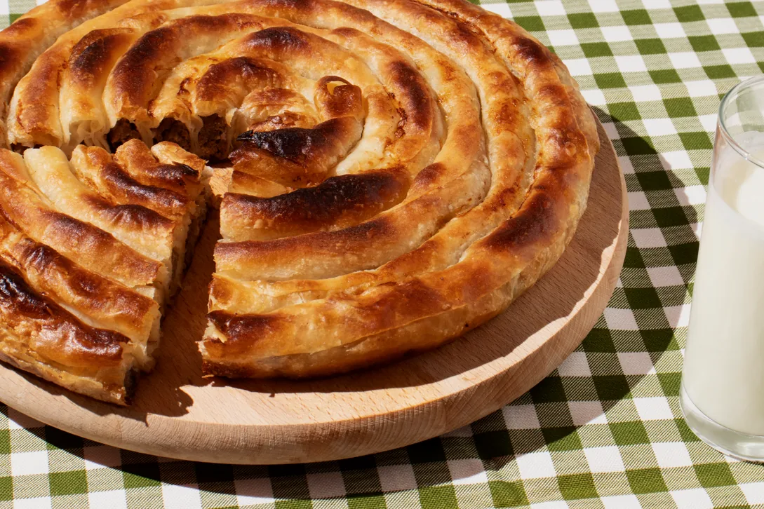 Golden spiral Bosnian beef burek with a cut wedge showing the meat filling, beside a glass of yogurt